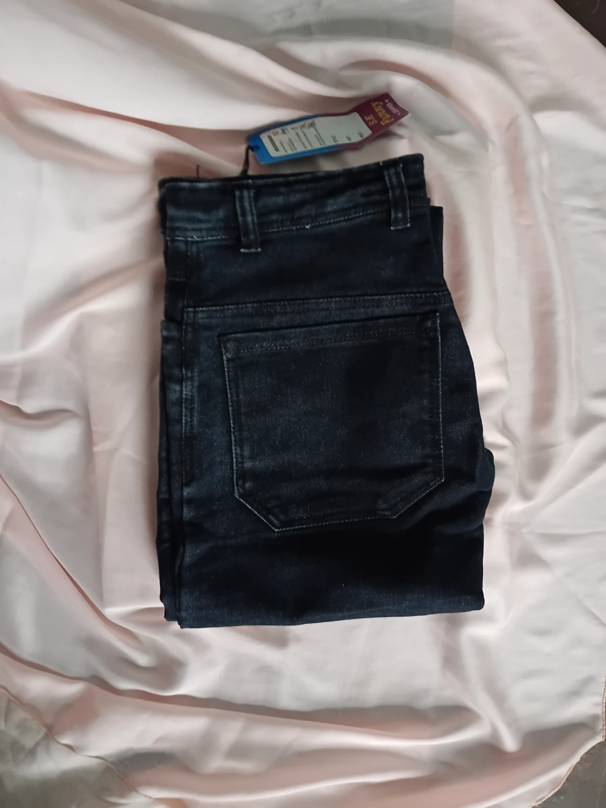 Folded dark wash denim jeans with back pocket detail and attached tag on a light fabric background