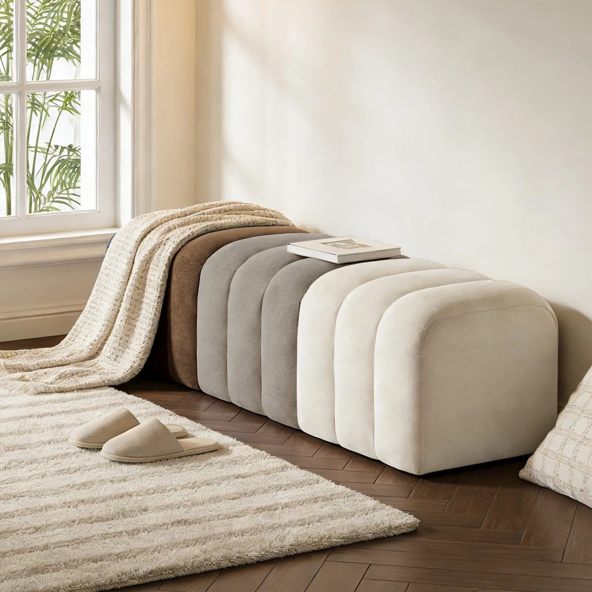 Side angle of the ribbed ottoman bench beside a window, styled with a throw blanket and book