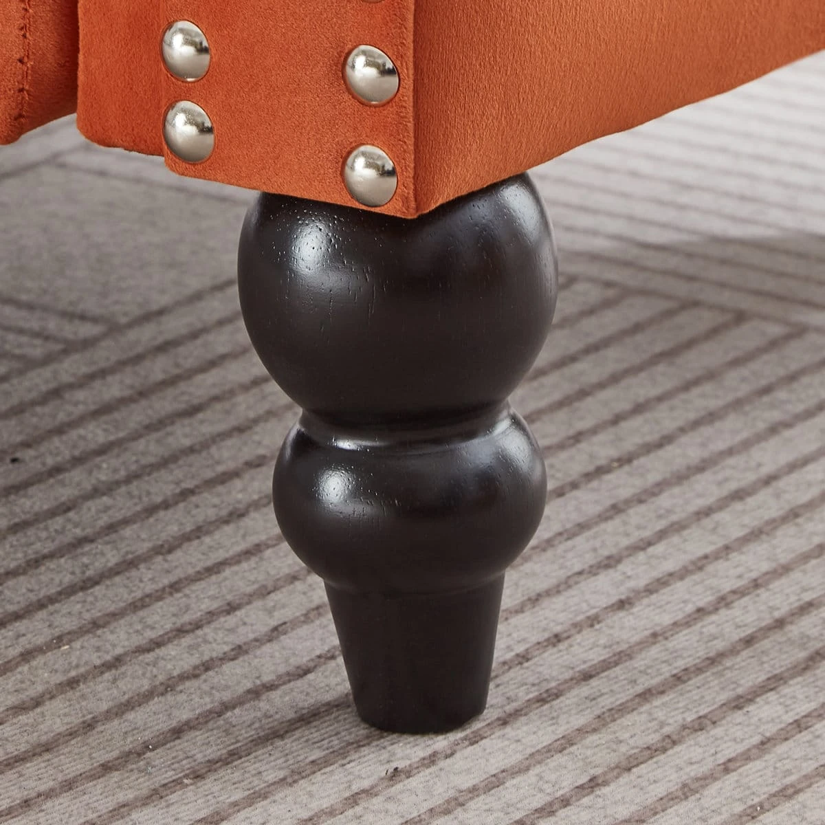 Close-up of dark turned wooden leg beneath orange upholstery with silver nailhead trim detail