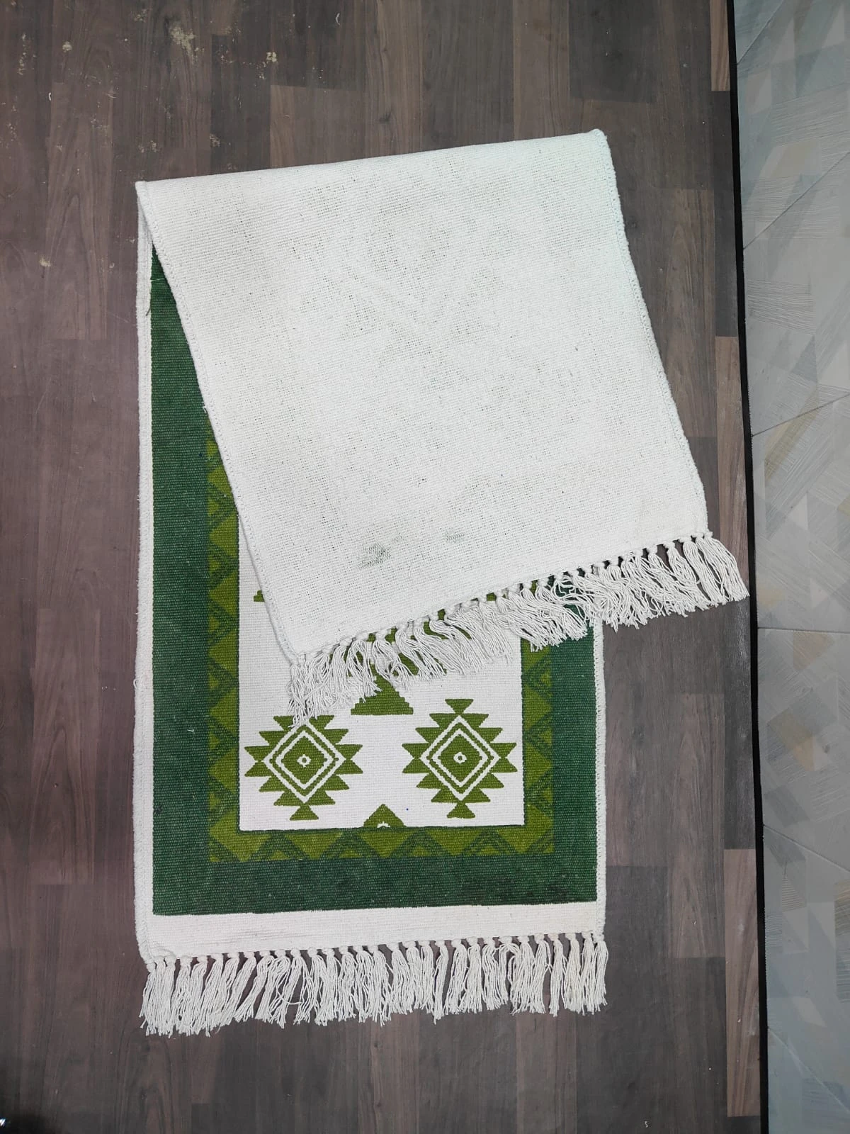 Partially folded runner rug showing plain cream reverse, tasseled edge, and green border with central diamond motifs