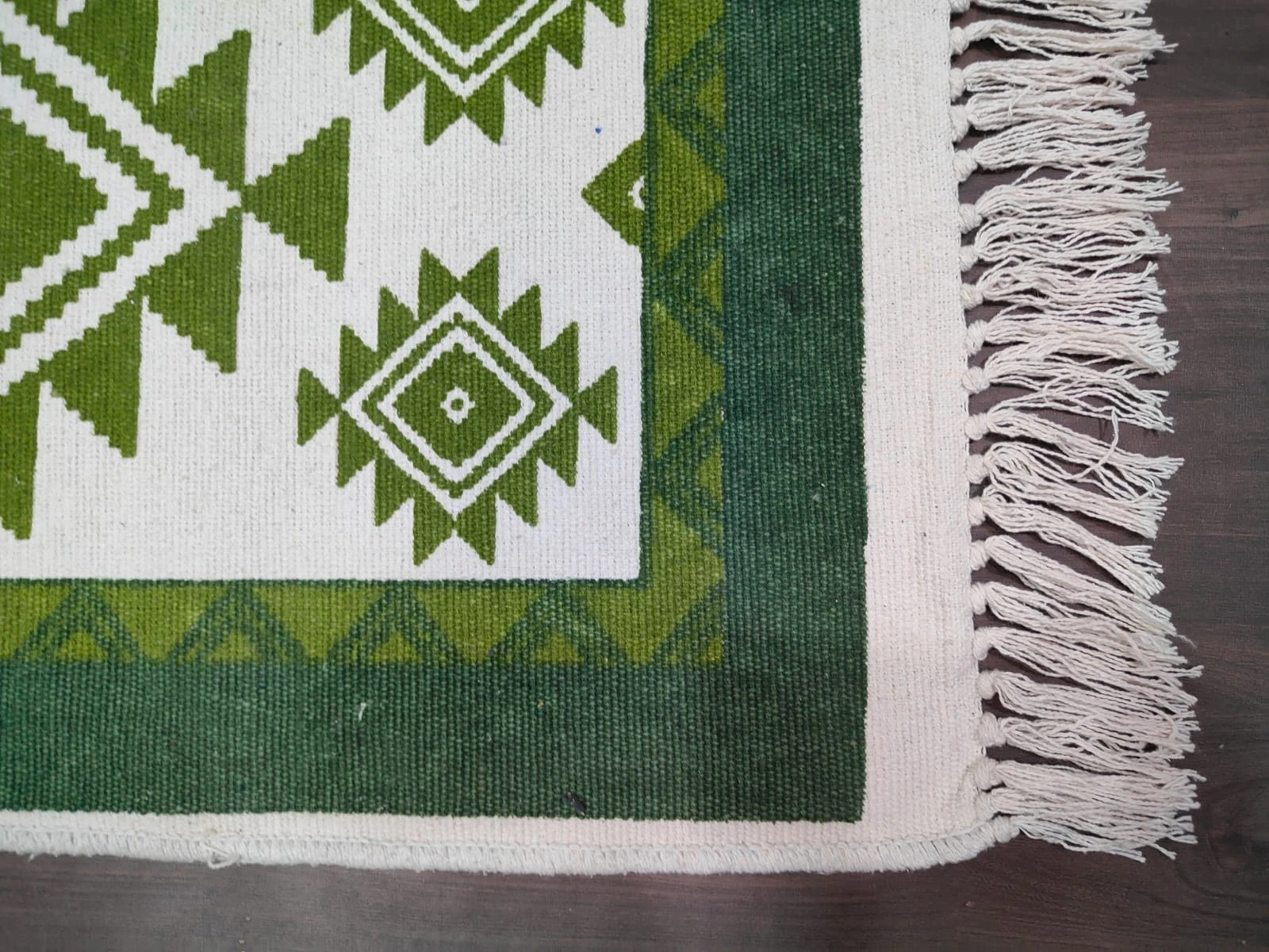 Close-up of rug corner with dark green border, cream edge binding, and white tassel fringe on one end