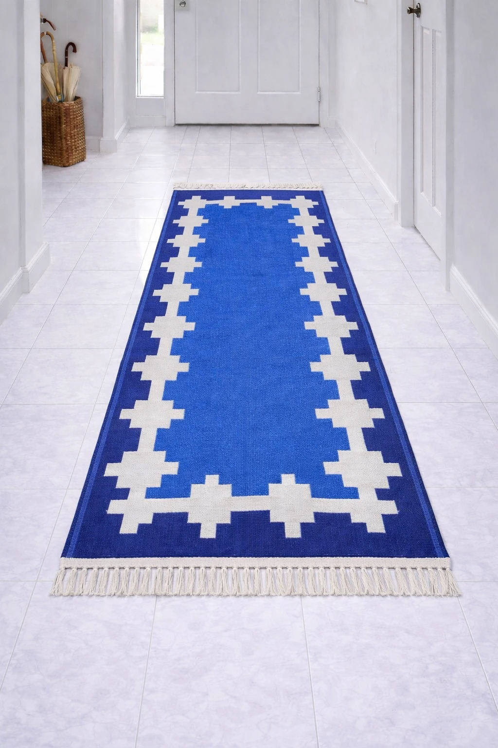 Blue runner rug with white stepped geometric frame and fringe, centered in a bright tiled hallway