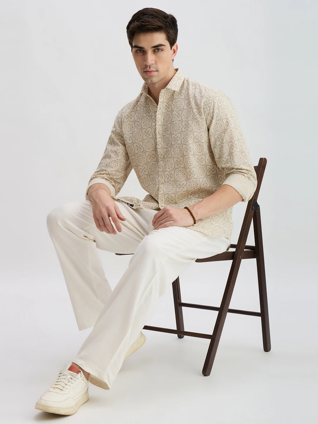 Seated full-body styling view showing the shirt worn with off-white trousers and rolled sleeves
