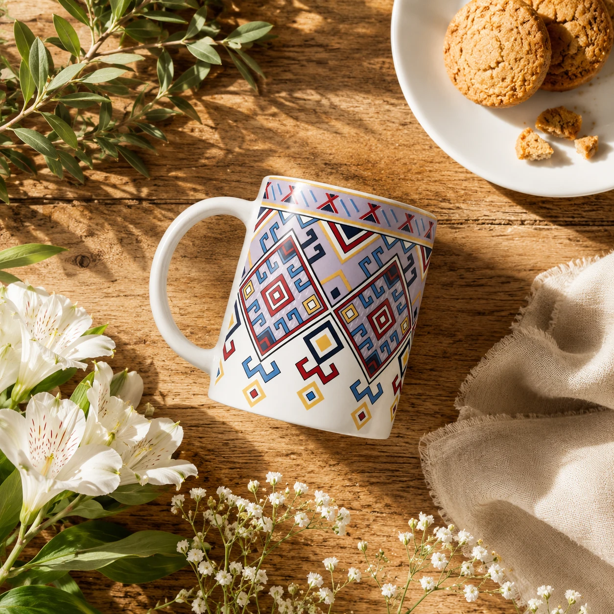 Geometric patterned white ceramic mug angled on a rustic table with cookies, flowers, and a beige cloth