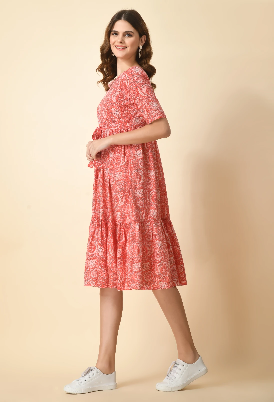 Side profile of coral floral midi dress with tie waist and tiered hem, paired with white sneakers