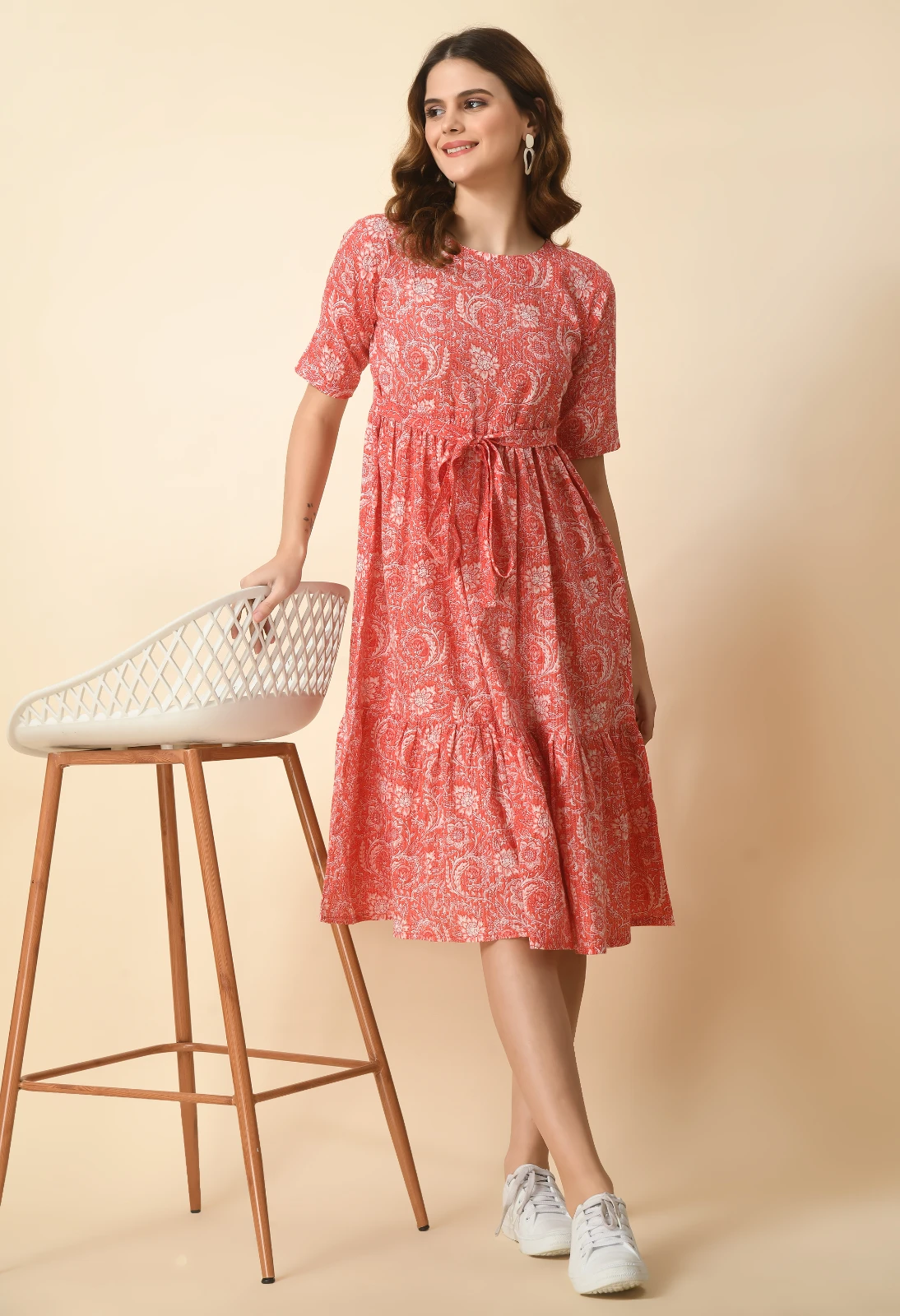 Three-quarter view of coral printed dress styled beside a white bar chair with wooden legs