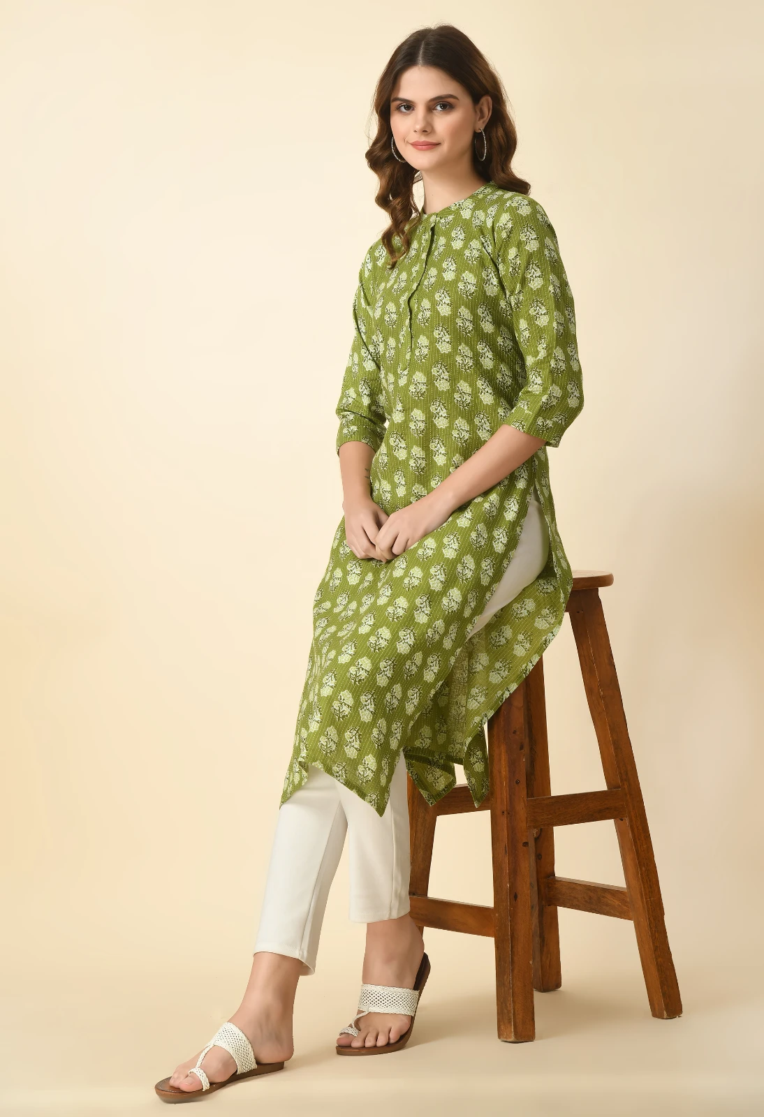 Seated angled view of olive green printed kurta with side slit, paired with off-white pants and flats