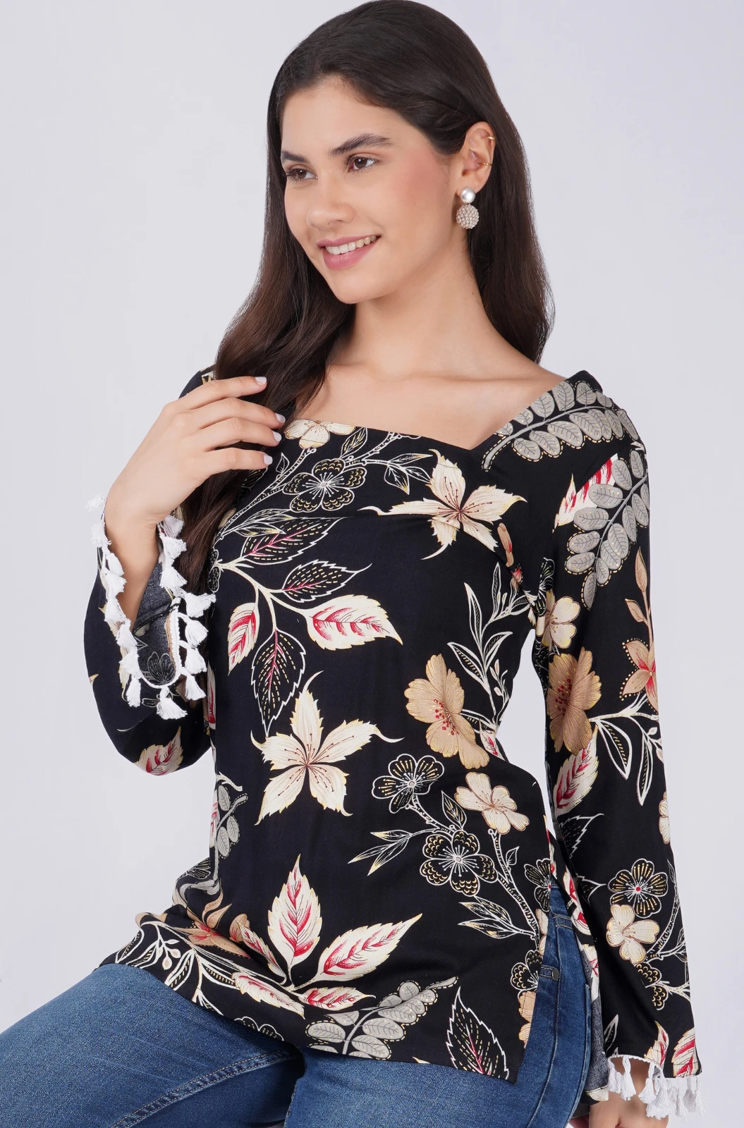 Three-quarter view of black floral top with tassel-trim bell sleeves, worn with jeans in a seated pose