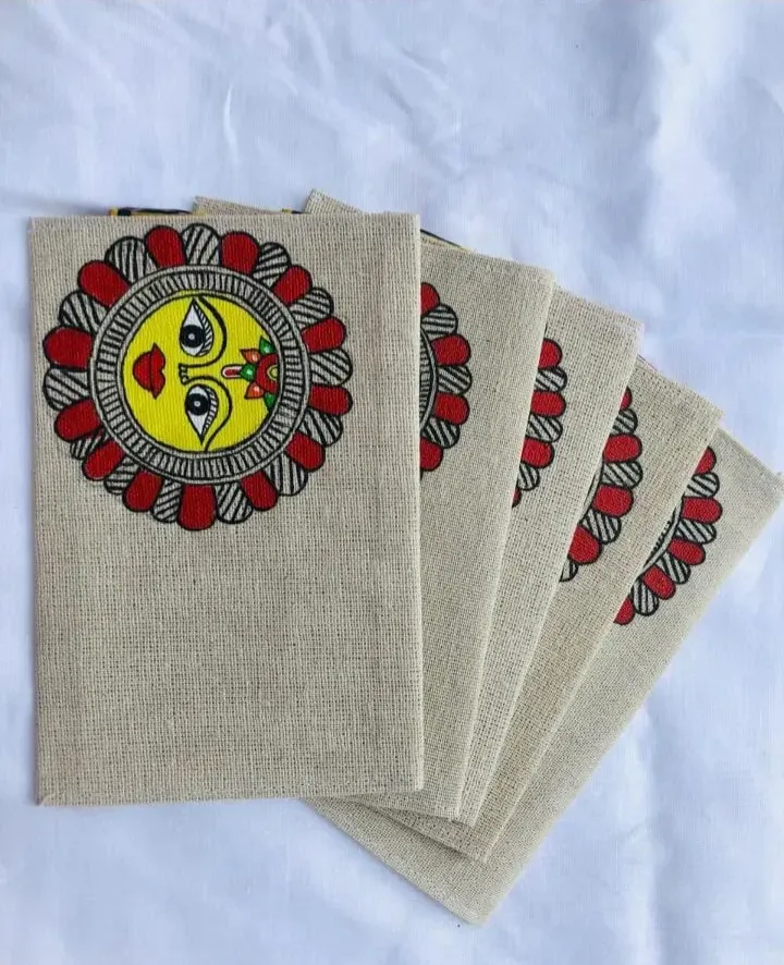 Four beige jute envelopes fanned out on white fabric, each with a colorful handpainted folk face motif