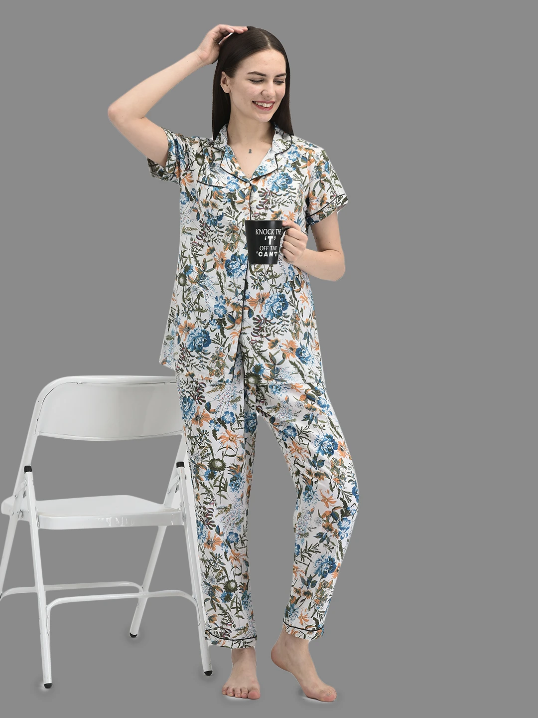 Model styling the floral pajama set while holding a black mug beside a white folding chair