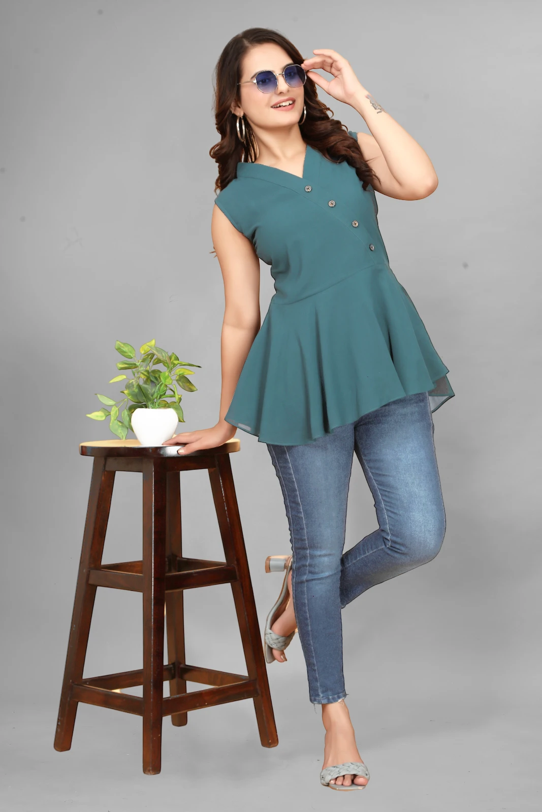 Woman wearing a teal sleeveless peplum top with front buttons, blue jeans and tinted sunglasses